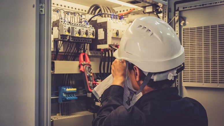 An electrician wearing a white hard hard and notepad inspecting the buildings electrical room.