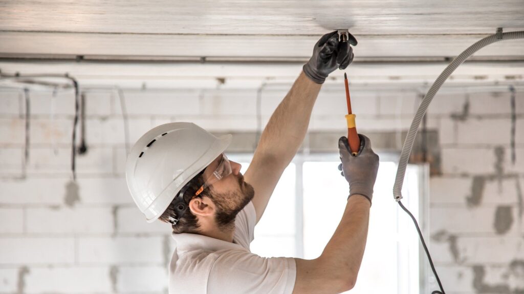 Electrician standing on a ladder in construction building that is using a screwdriver to screw in bolts